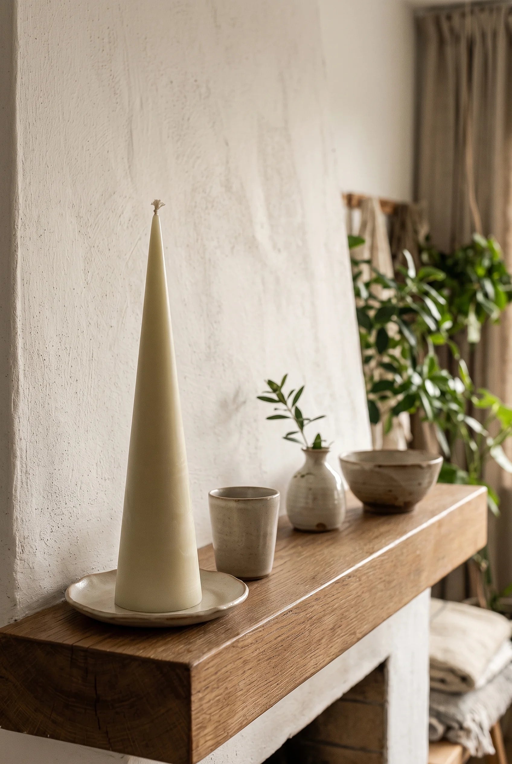 Et højt keglelys i råhvid bivoks på et lille wabi-sabi keramikfad på kaminhylden, ved siden af en lille vase med en grøn kvist og en stentøjsskål.