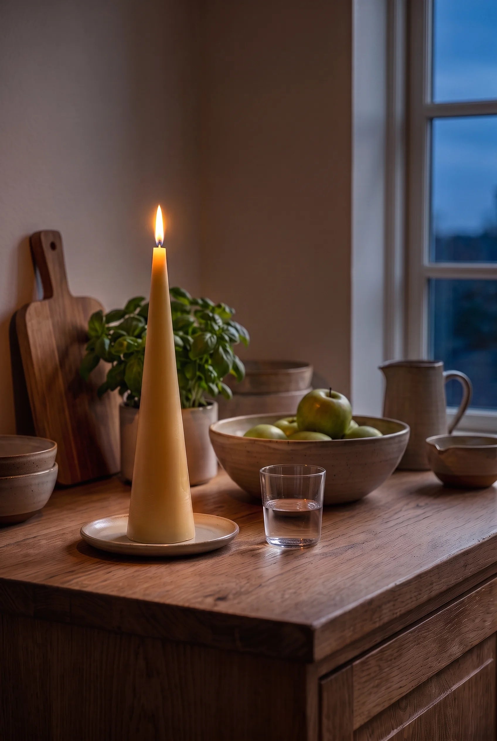 Et tændt gyldent keglelys på køkkenøen om aftenen — flammen trækker et lunt skær over æbleskålen i det mørke køkken.