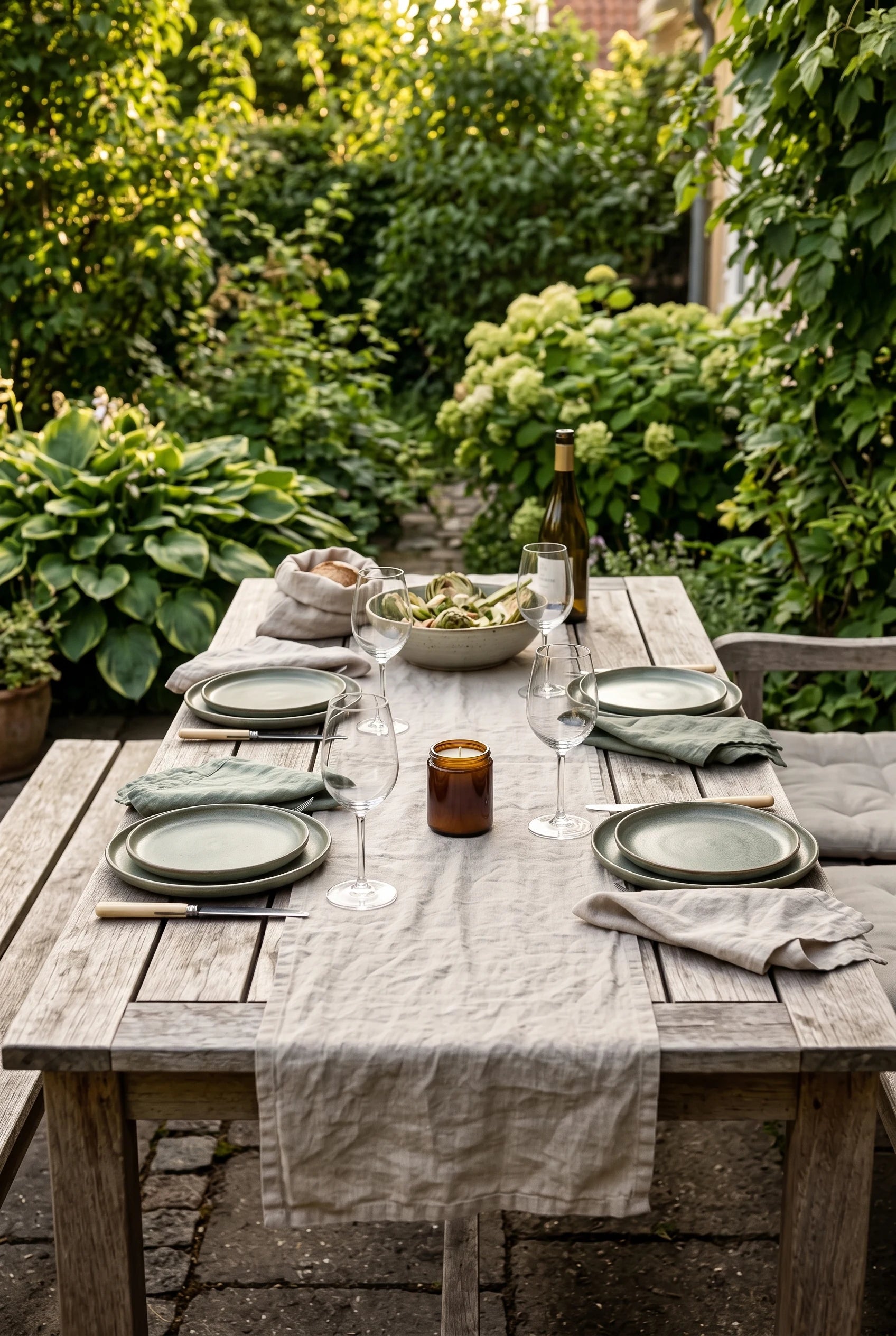 Et langt havebord dækket til middag i aftensolen — grønne stentøjstallerkener, hvidvin og artiskoksalat, med Reden bivokslys i brun krukke midt på bordet, klar til at blive tændt.