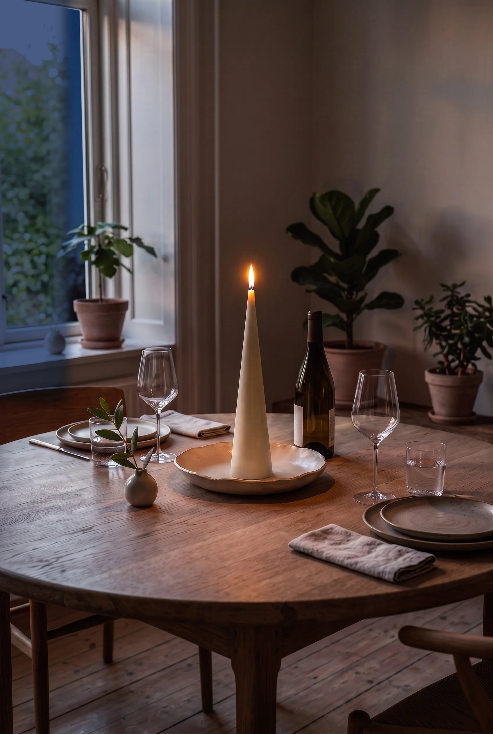 Et tændt keglelys i råhvid bivoks midt på et bord dækket til to om aftenen — flammen på keglens spids er bordets eneste lys.