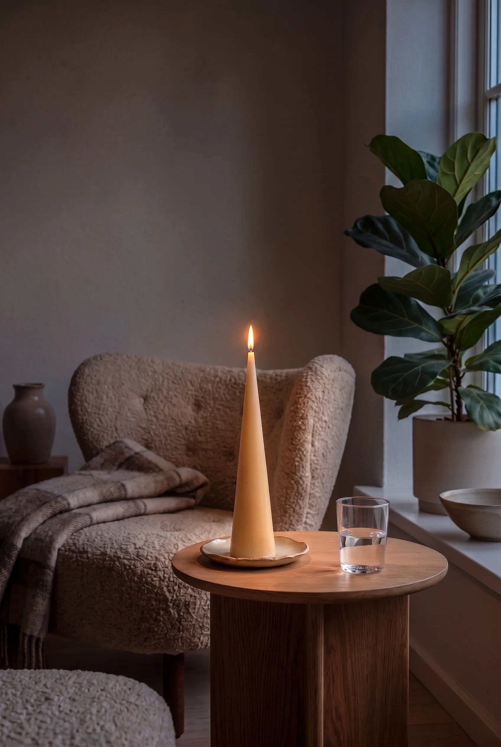 Et tændt Grankoglen keglelys ved lænestolen om aftenen — en lille afdæmpet lampe gløder bagved, og flammen gør læsekrogen til aftenens bedste plads.