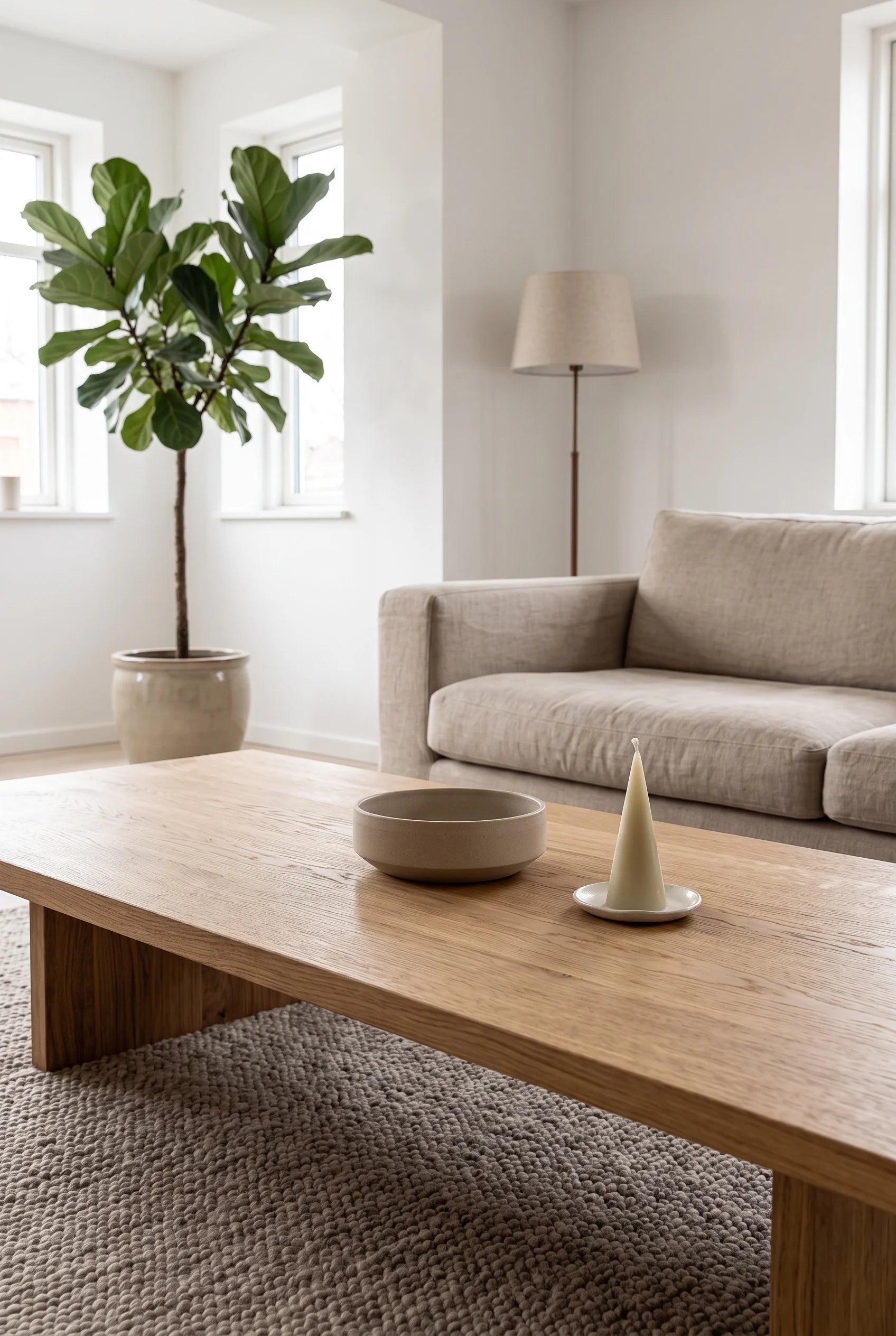 En rolig dansk stue med sofa i sandfarvet hør og stort egetræssofabord — et lille keglelys i råhvid bivoks på sit keramikfad ved siden af en lav rund stentøjsskål.