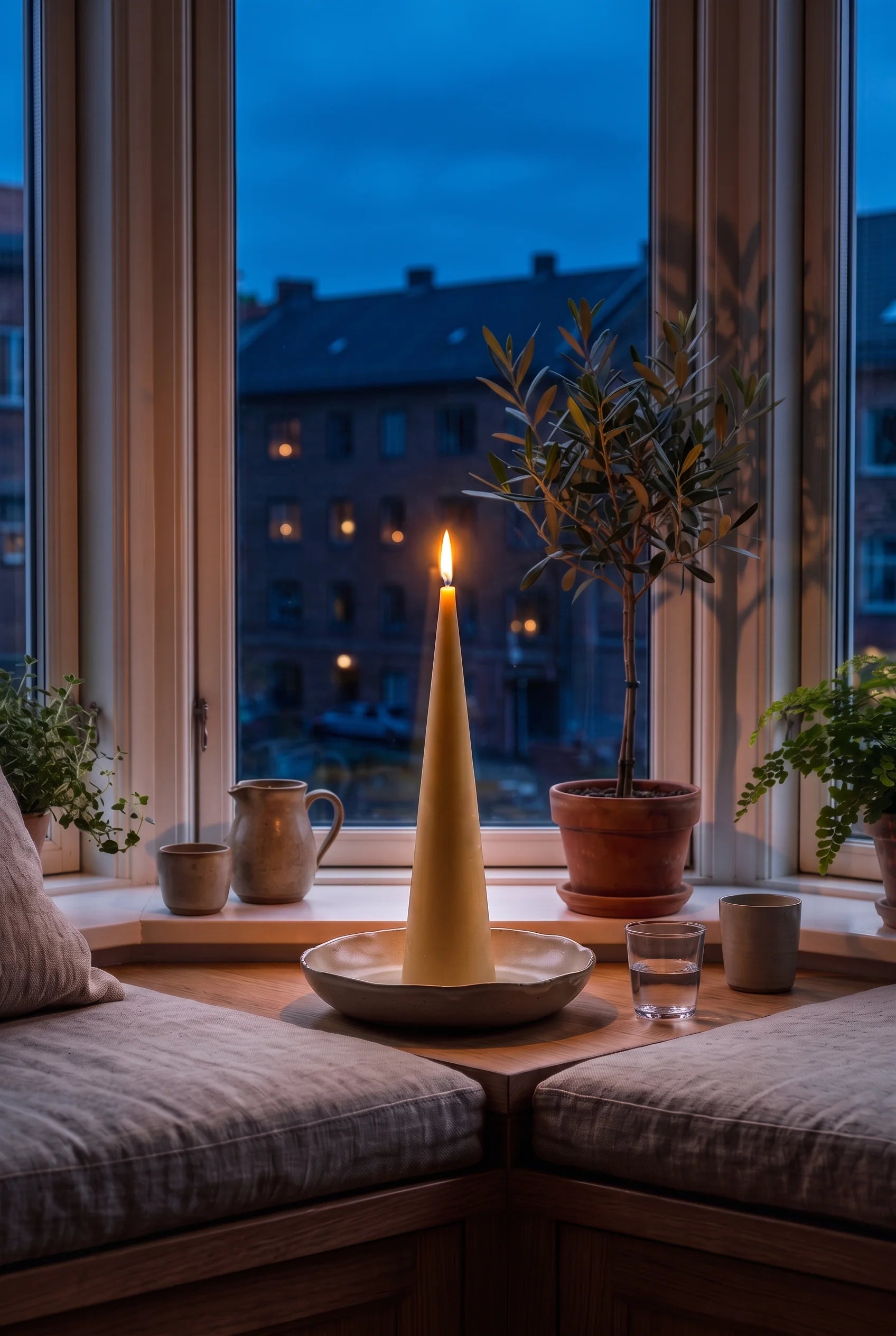Et tændt Grankoglen keglelys i karnappen om aftenen — flammen står roligt mod de oplyste vinduer på den anden side af gaden.