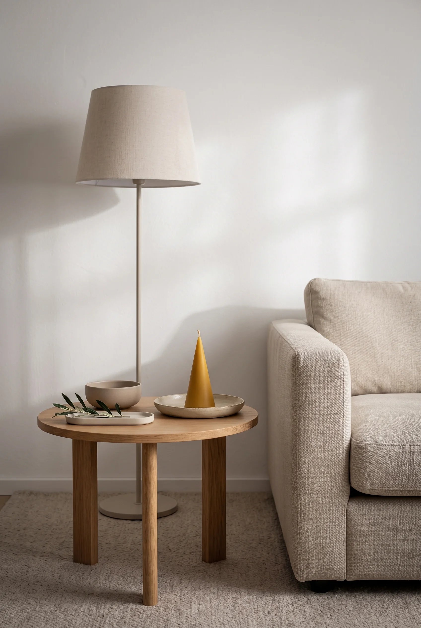 En rolig dansk stue med sandfarvet hørsofa og rundt egetræssidebord — et lille keglelys i gylden bivoks på sit fad, med en gulvlampe med stofskærm bagved.