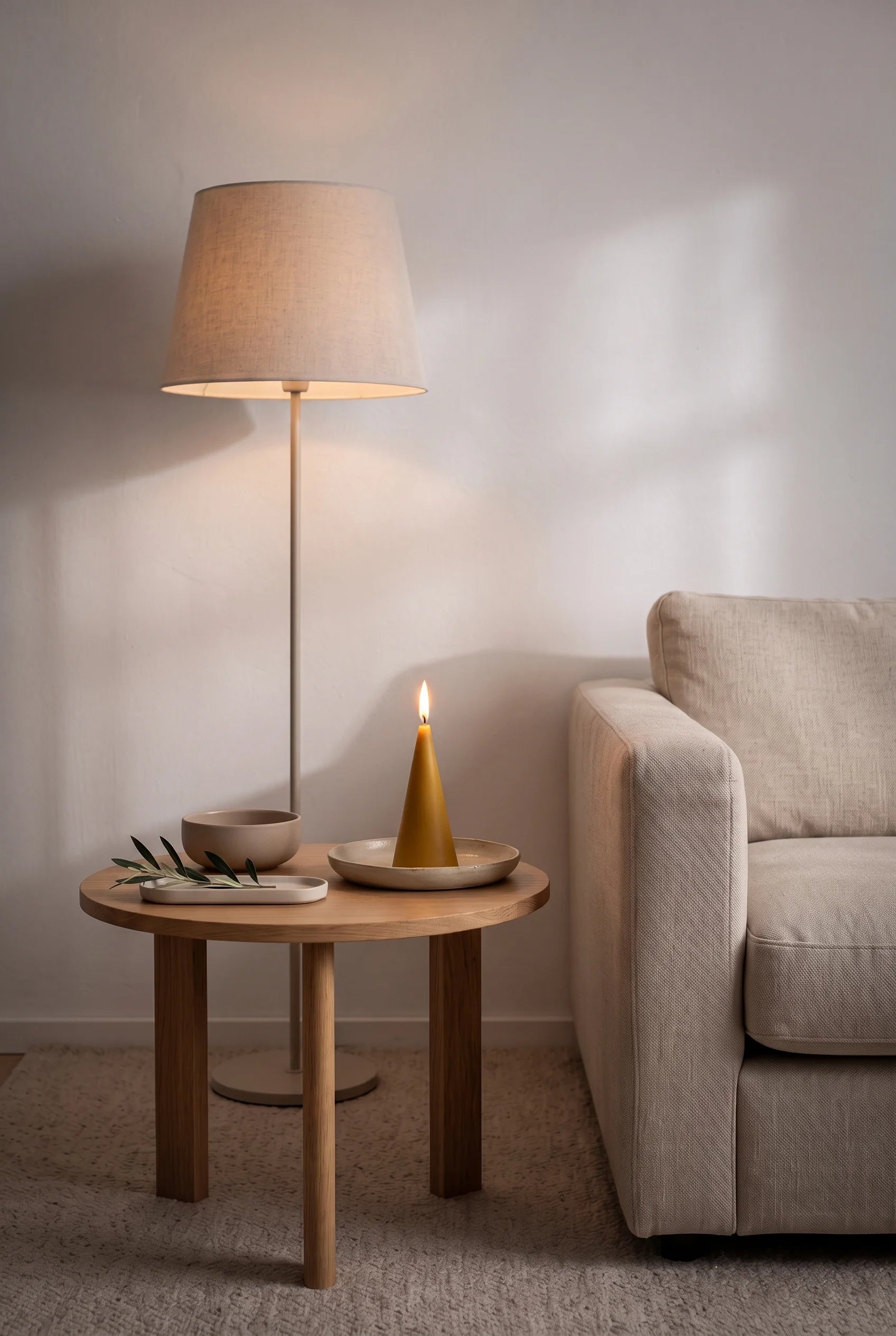 Et tændt keglelys i gylden bivoks på sidebordet ved sofaen om aftenen — gulvlampen lyser afdæmpet, og flammen samler stuen i et blødt, varmt skær.